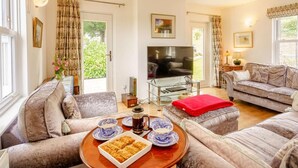 A living room with a TV, sofas, a coffee table with a tray of food, and a view of greenery outside. - 5 Bed in Askam-in-furness (oc-89226) (Kirkby Ireleth)