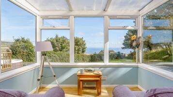 A conservatory with a view of the sea, a wooden table with a lamp, and two purple sofas.