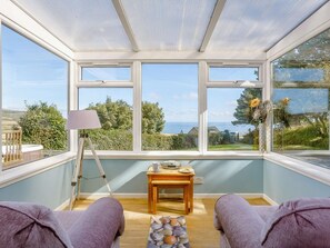 Ein Wintergarten mit Meerblick, ein Holztisch mit Lampe und zwei violette Sofas.