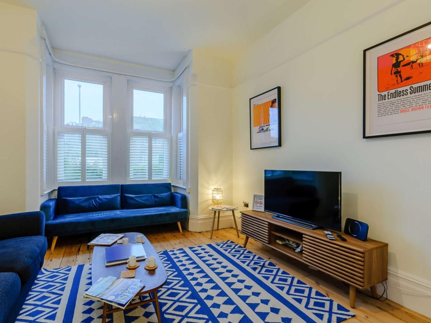 Ein Wohnzimmer mit einem blauen Sofa, einem Fernseher auf einem Holzgestell, einem gemusterten Teppich und einem Fenster mit Jalousien.