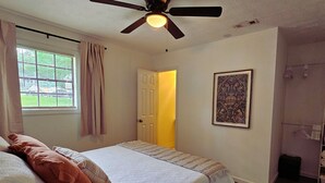 Ein Schlafzimmer mit einem Bett, einem Deckenventilator, einem Fenster mit Vorhängen, einer Tür zu einem anderen Raum und einem gerahmten Bild an der Wand.