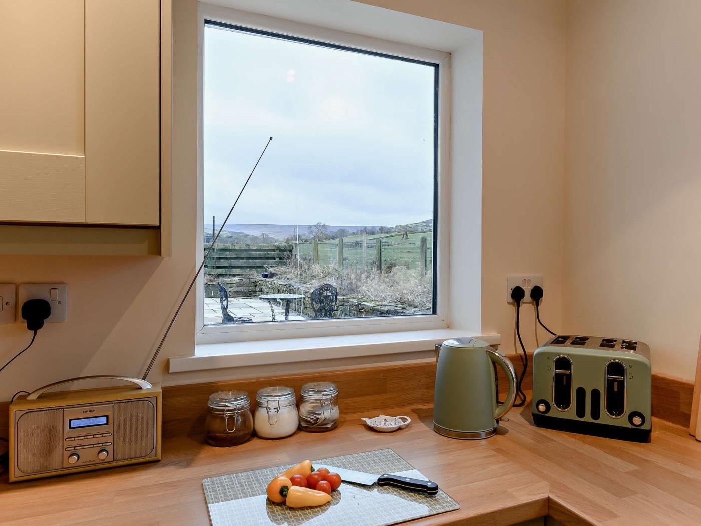 A kitchen with a window, a radio, jars, a kettle, a toaster, and a cutting board with vegetables.