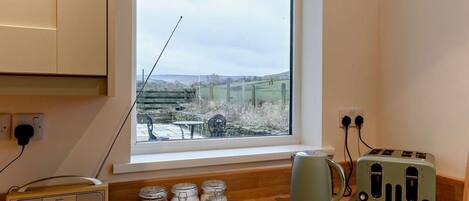 A kitchen with a window, a radio, jars, a kettle, a toaster, and a cutting board with vegetables.