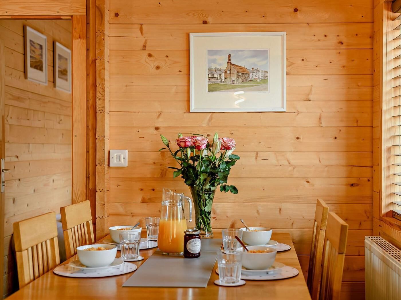 Une table de salle à manger en bois, ornée d’un vase de fleurs, d’une carafe de jus d’orange et de bols de céréales.
