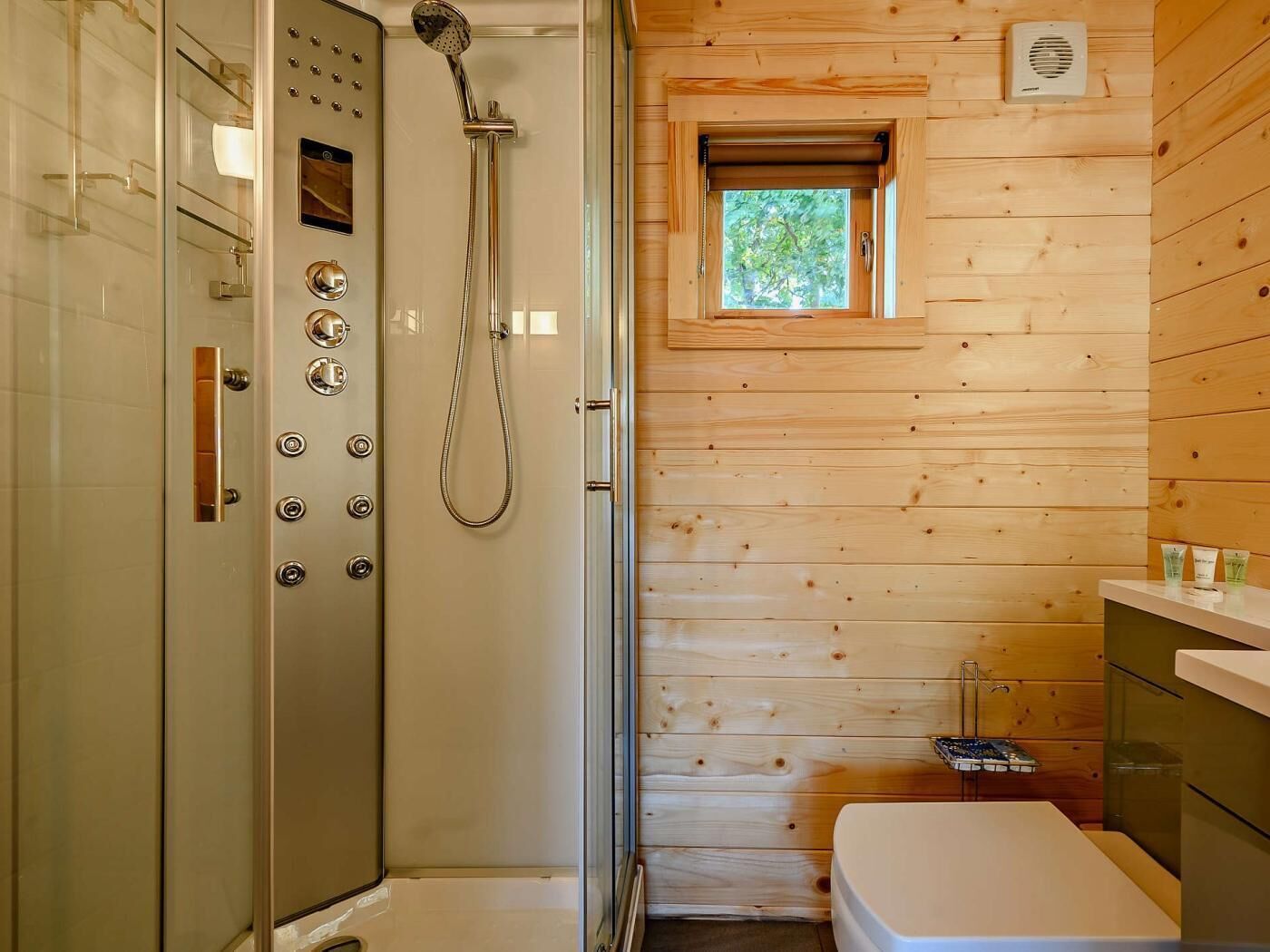 Une salle de bain rustique, avec une cabine en bois, une douche fermée par des parois de verre, une petite fenêtre et un lavabo blanc.