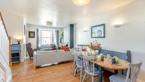 A modern living room with a sofa, dining table, and chairs, a staircase, and a large window with curtains. - 3 Bed in Bideford (oc-89991) (Appledore)