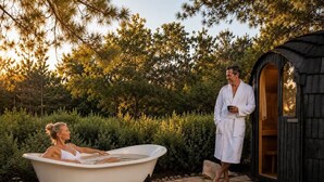 A woman relaxing in a bathtub outdoors, a man standing nearby, and a small wooden structure.