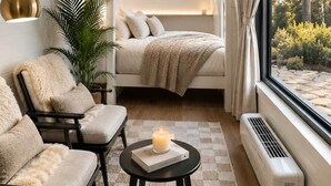 A cozy room with a bed, two chairs, a small table with a candle, and a large window with a view of trees.
