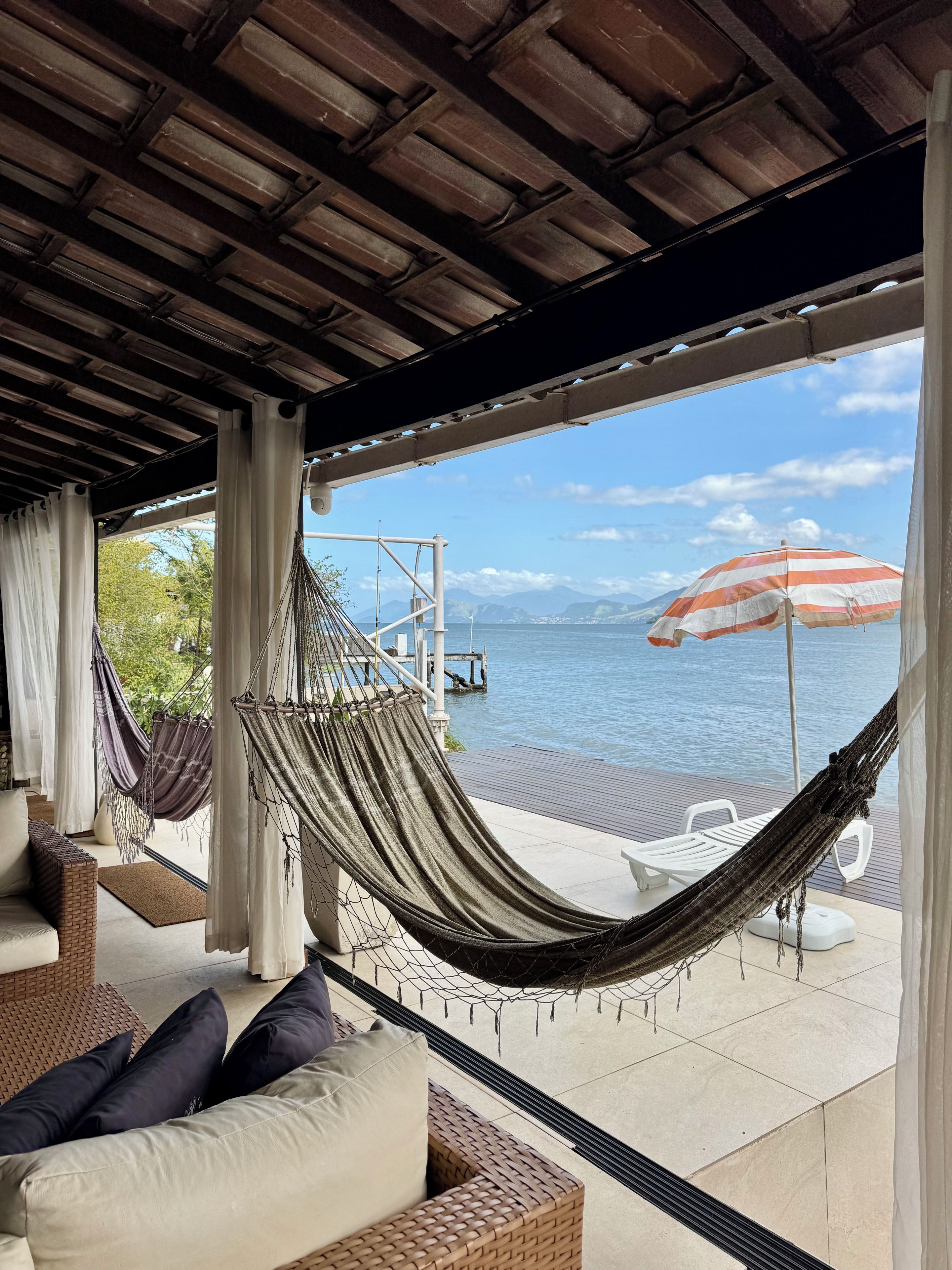 Eine Hängematte unter einer Holzbalkendecke, ein Korbsessel mit Kissen und Blick aufs Meer mit Sonnenschirm.