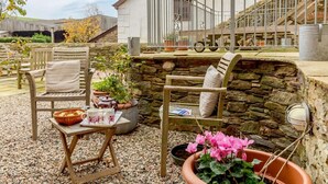 Un patio con sillas de madera, una mesa con una cesta y tazas, plantas en macetas y un muro de piedra.
