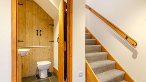Un baño con puerta de madera, un lavamanos blanco y un inodoro. Se observa una escalera con barandillas de madera y peldaños alfombrados.