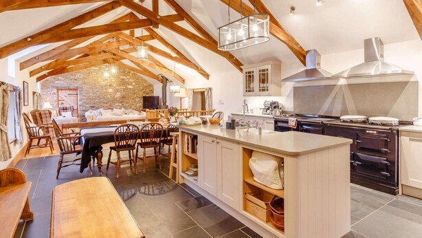 A spacious kitchen with a central island, wooden beams, and a stone wall in the background. - 4 Bed in Kingsbridge (oc-90027) (South Huish)