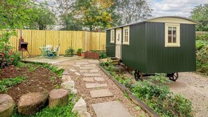 Una casa móvil verde pequeña con una ventana, una cerca de madera, un jardín con piedras para pisar y una parrilla.