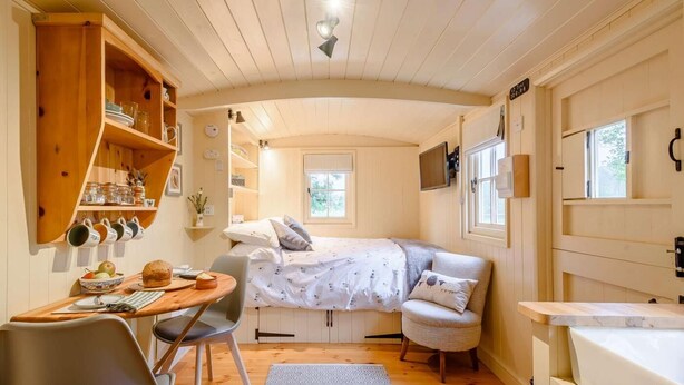 Una habitación acogedora con una cama, una mesa de comedor con sillas, un estante de madera con vajilla y una ventana.