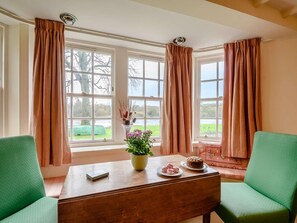 A room with a wooden table, two green chairs, a vase with flowers, and a view of a lake through large windows with curtains. - 4 Bed in Ayr- (oc-90038) (Ayr)