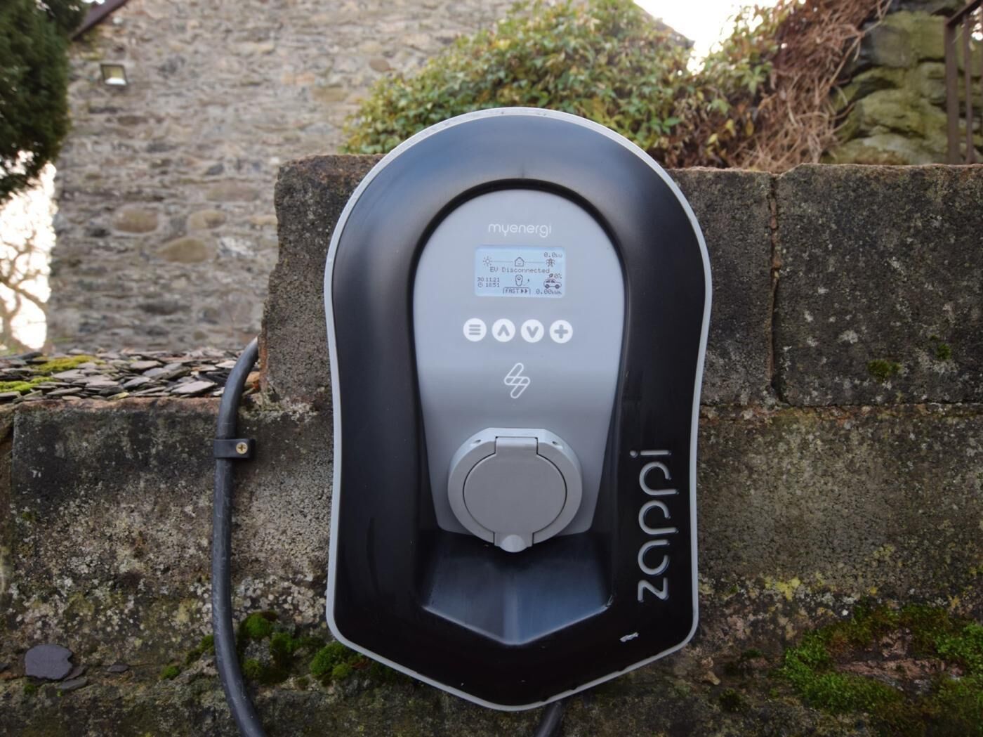 An electric vehicle charging station branded 'myenergi' with a cable connected to it, mounted on a stone wall.