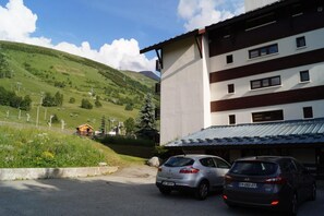 Ein Parkplatz mit zwei Autos, ein Gebäude mit einem Satteldach und ein grüner Hang im Hintergrund.