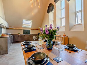 Una cocina moderna con una larga mesa de comedor dispuesta para una comida, un florero con flores y vistas a la ciudad a través de amplias ventanas.