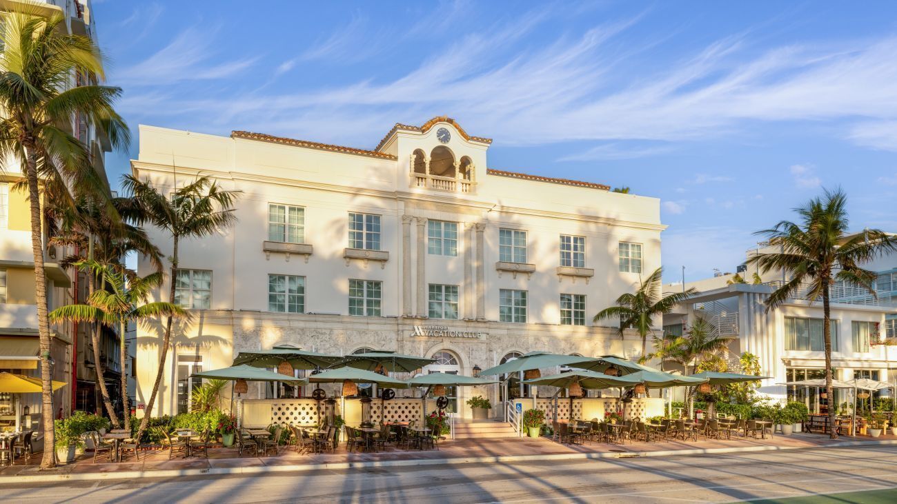A hotel with a central entrance, outdoor seating area with umbrellas, and palm trees.