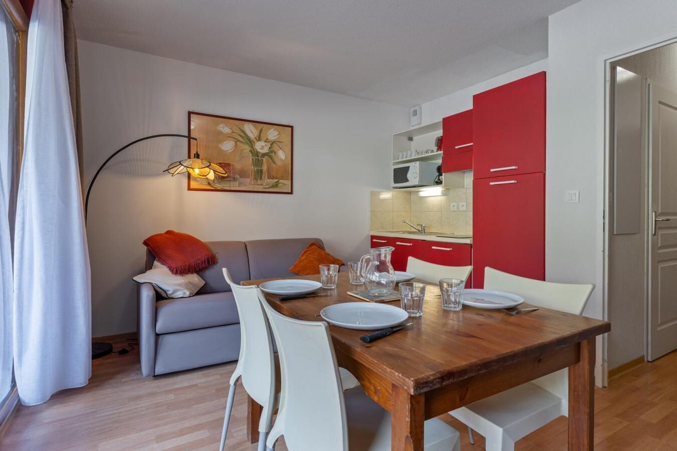 Ein Essbereich mit einem Holztisch, gedeckt für vier Personen, ein graues Sofa mit roter Decke sowie eine Küche mit roten Schränken.