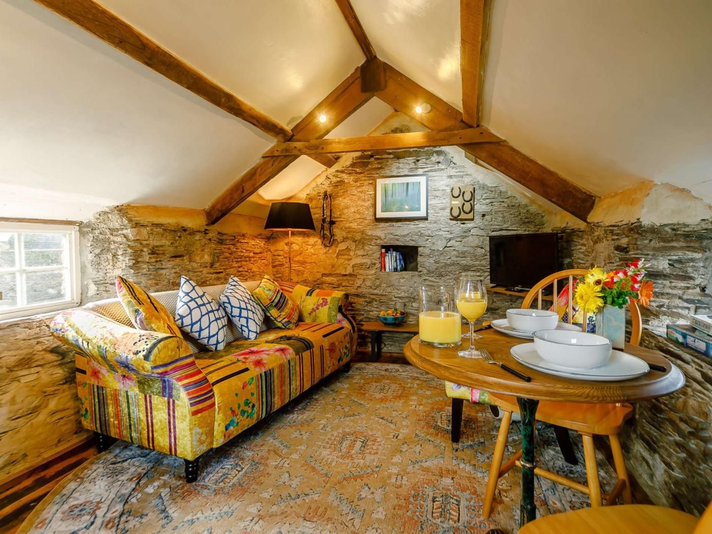 A cozy loft-style room with a stone wall, a colorful sofa, a dining table with chairs, and a television mounted on the wall.
