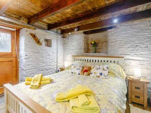 A rustic bedroom with a wooden ceiling, stone walls, a bed with floral bedding, a teddy bear, and a nightstand with a lamp. - 1 Bed in Saltash (oc-90072) (Trerulefoot)