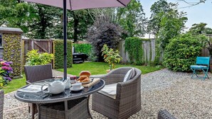 Eine Terrasse im Freien mit Korbmöbeln, einem Tisch gedeckt mit einem Teekessel, Tassen und Croissants, sowie einem violetten Sonnenschirm, der Schatten spendet.