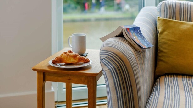 Ein gemütliches Zimmer mit einem gestreiften Sofa, einem kleinen Holztisch mit einem Croissant, einer weißen Tasse und einem Buch.