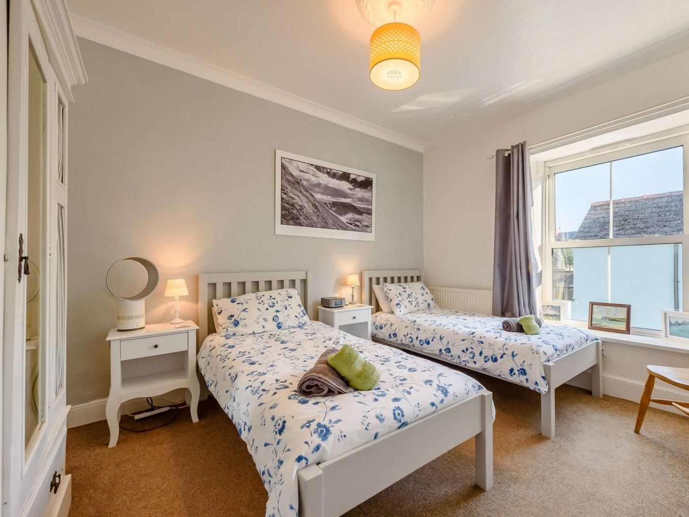 A bedroom with two beds, bedside tables, a lamp, a picture on the wall, and a window with curtains.