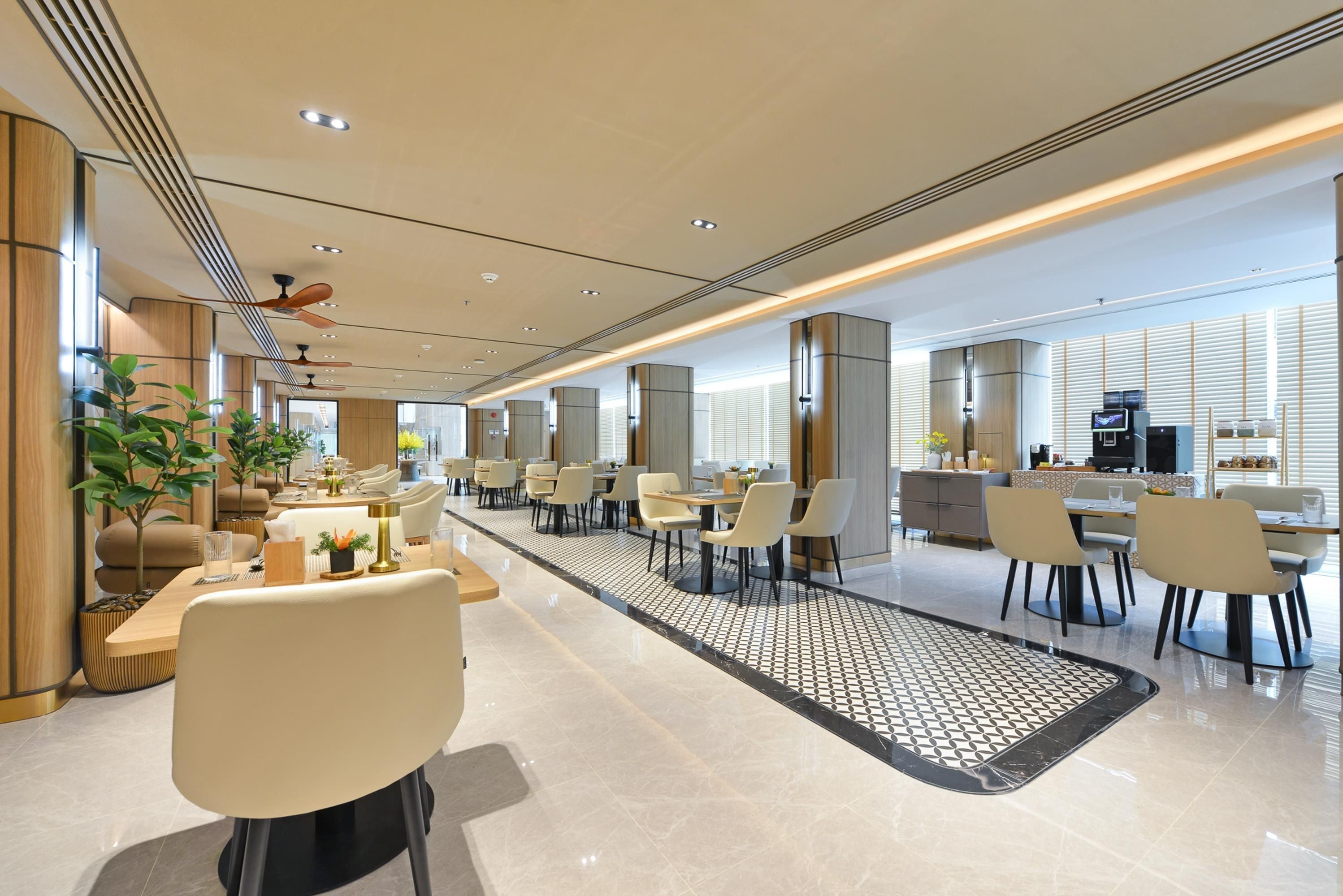 Espaço de refeições moderno em hotel, com várias mesas, cadeiras e um corredor central, além de área de cozinha ao fundo.