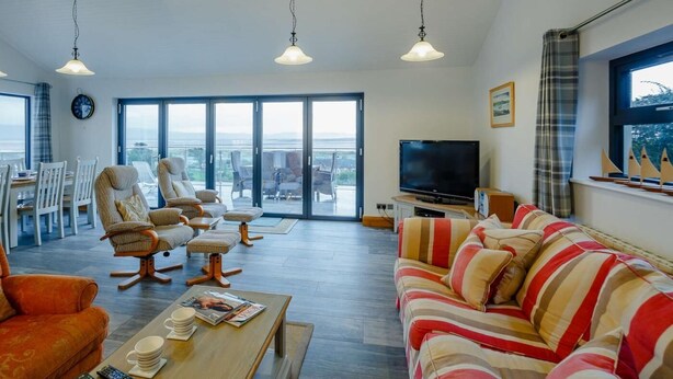 A spacious living room with a large TV, comfortable seating, a dining area, and a balcony with a view. - 5 Bed in Aberdovey (oc-90222) (Aberdovey)