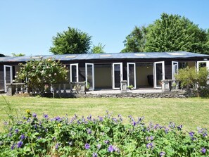 Un bâtiment à deux étages doté d’un patio couvert, de marches en pierre menant à l’entrée et d’un jardin où l’on peut admirer des fleurs violettes au premier plan.