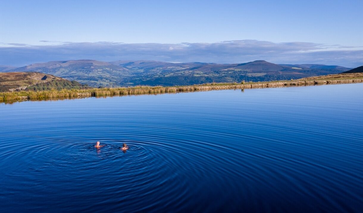 A calm lake with two people swimming, surrounded by hills and clear skies.