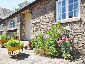 Una cabaña de piedra con un letrero que dice 'Granary Cottage', un carretón amarillo y plantas en macetas.