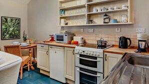 A kitchen with white cabinets, a microwave, a stove, and a sink. There are shelves above the counter holding cups and glasses. - 1 Bed in Kirkby Stephen (oc-89457) (Crosby Garrett)