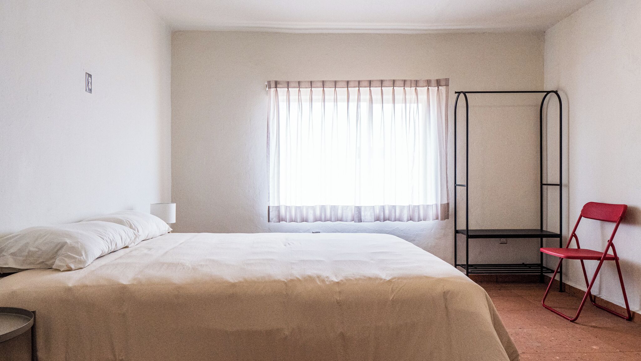 Una habitación con una cama, una ventana con cortinas translúcidas, una silla roja y un estante metálico.