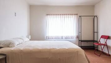 Una habitación con una cama, una ventana con cortinas translúcidas, una silla roja y un estante metálico.
