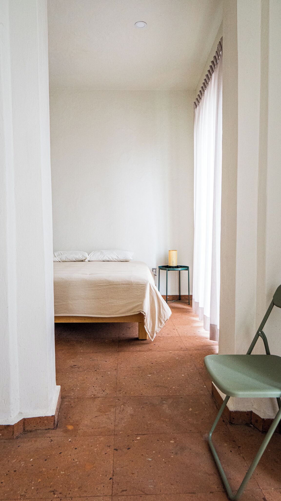 Una habitación con cama, una silla, una mesita de noche con lámpara y una ventana con cortinas.
