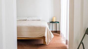 Una habitación con cama, una silla, una mesita de noche con lámpara y una ventana con cortinas.