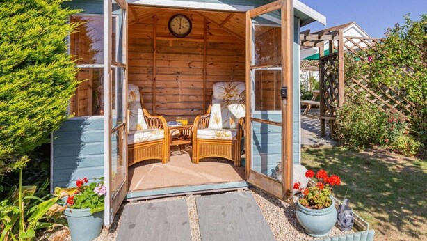Eine Holzhütte mit offener Vorderseite, zwei Korbstühle, ein Tisch und eine Uhr an der Wand.