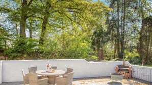 Een terras buiten met rieten meubilair, een ronde tafel met een bloevas en een barbecue.