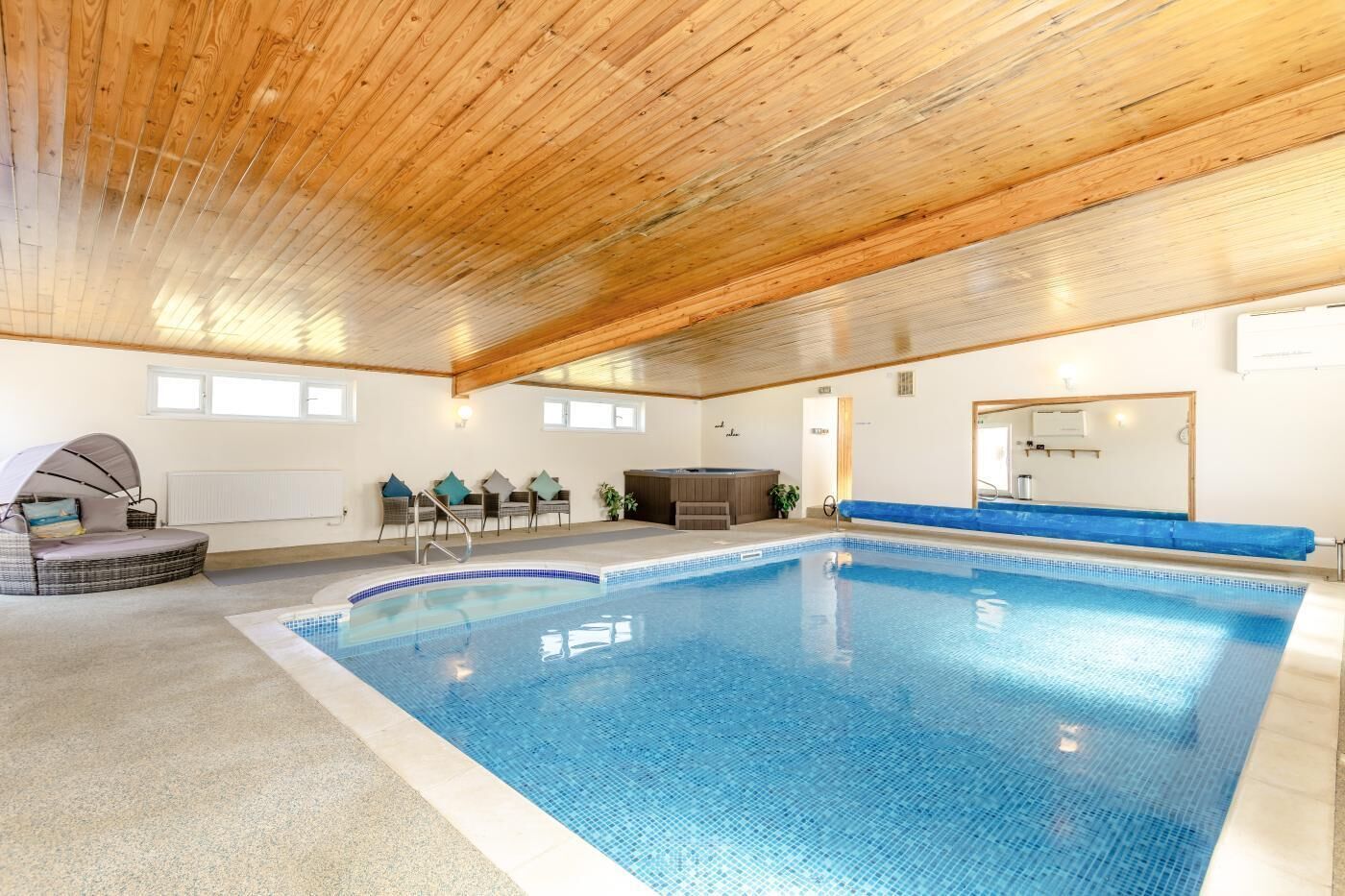 Una piscina con borde de azulejos, techo de madera y un área de descanso con sillas y un banco acolchado.