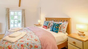 Ein Schlafzimmer mit einem hölzernen Bett, blumigem Bettzeug, einem Nachttisch mit Lampe und einem Fenster mit Vorhängen.