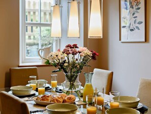 Ein Esstisch ist gedeckt mit Tellern, Gläsern und einer Blumenvase. Im Hintergrund ist ein Fenster und Kunstwerk zu sehen.