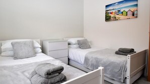 Un dormitorio con dos camas, ropa de cama blanca, una mesita de noche, una cómoda y un cuadro con motivos playeros en la pared.