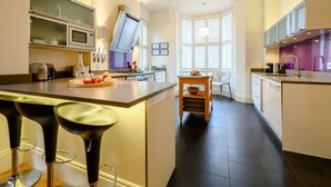 A modern kitchen with a central island, bar stools, a dining area, and a window with blinds. - 4 Bed in Keswick (oc-89258) (Keswick)