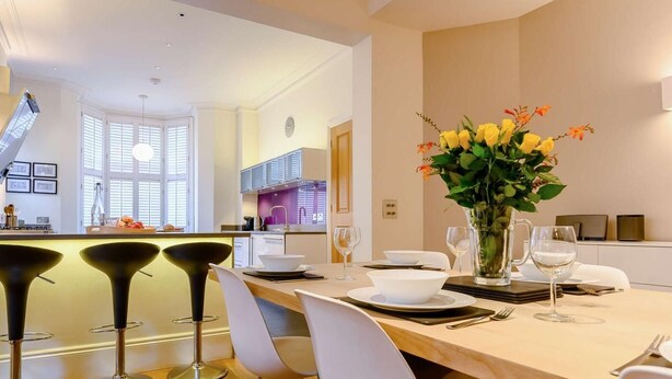 A modern kitchen with a central island, a dining area with bar stools, and a vase of flowers on the table. - 4 Bed in Keswick (oc-89258) (Keswick)