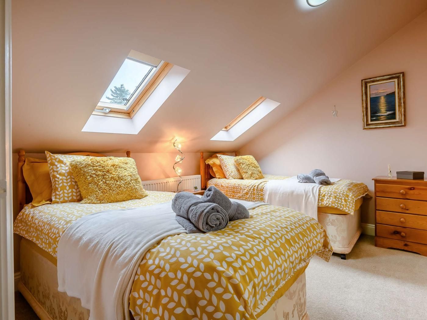 A bedroom with two beds, a skylight, a bedside lamp, a dresser, and a framed picture on the wall.
