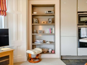 A modern kitchen with a built-in oven, a wooden shelf unit with books and decorative items, and a unique stool with a cushioned seat. - 2 Bed in Keswick (oc-89261) (Portinscale)