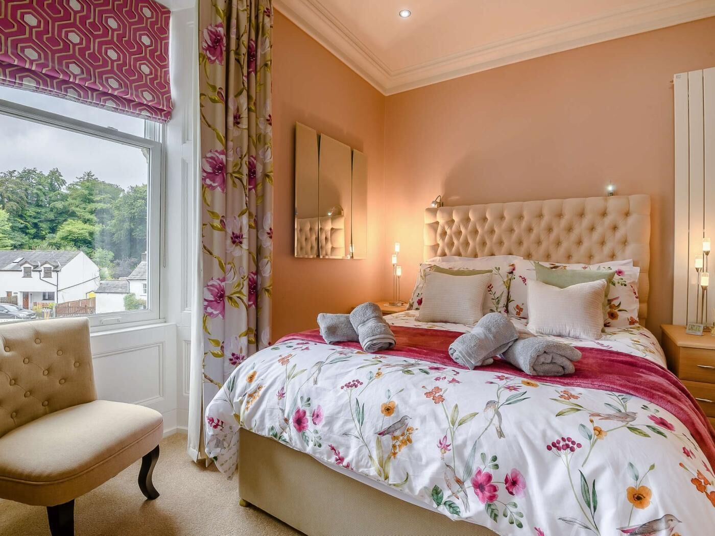 A bedroom with a large bed, a chair, a window with curtains, and a view of houses outside.